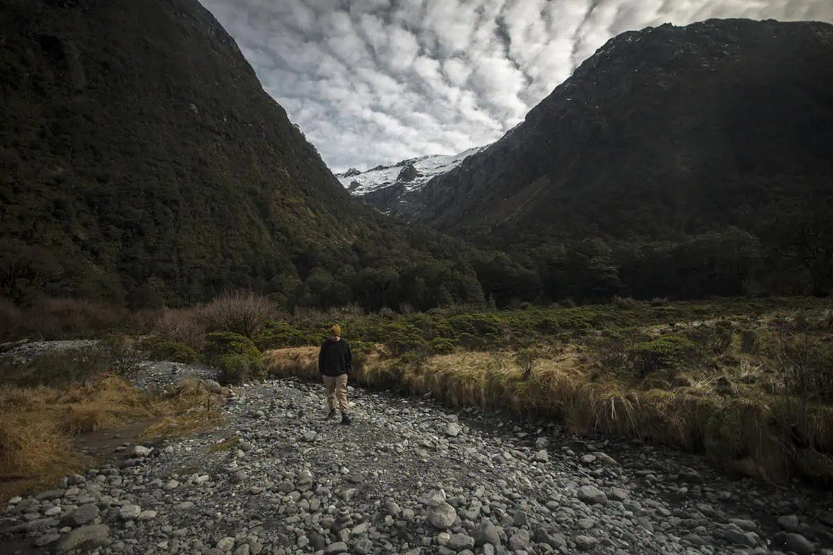Monkey Creek: BEST Stop On The Way To Milford Sound 14 A lone hiker walks along a rocky path at Monkey Creek, New Zealand, surrounded by a dense forest and steep, snow-capped mountains. The person, wearing a dark jacket and yellow beanie, appears small against the vast, dramatic landscape. The sky above is filled with clouds, adding to the sense of solitude and wilderness.