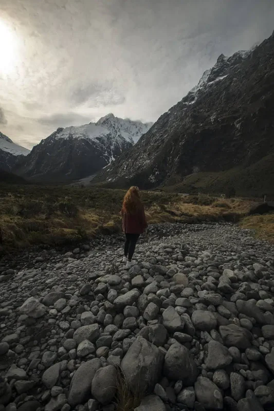 Monkey Creek: BEST Stop On The Way To Milford Sound 22 tasha amy with long, red hair walks along a rocky streambed at Monkey Creek, New Zealand. She is dressed in a rust-colored sweater and black pants, and the rugged mountains rise dramatically behind her. The overcast sky and misty atmosphere enhance the wild, untamed beauty of the landscape, emphasizing the grandeur of the natural setting.