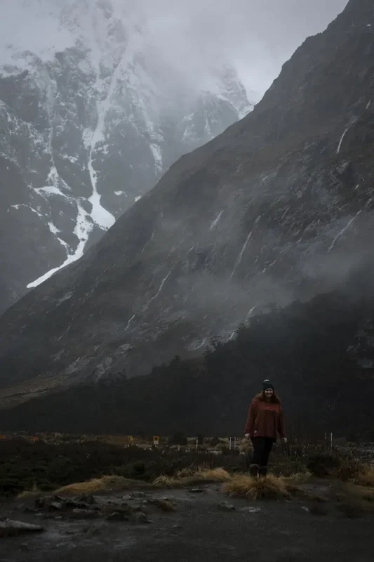 Monkey Creek: BEST Stop On The Way To Milford Sound 10 tasha amy dressed in a rust-colored sweater, black pants, and a beanie stands in the foreground of Monkey Creek. She is surrounded by rugged terrain and mist-covered mountains that rise sharply behind her. The dark, overcast sky and low clouds enhance the wild and untamed beauty of the landscape.
