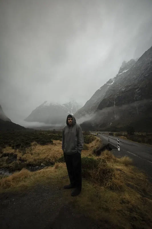 Monkey Creek: BEST Stop On The Way To Milford Sound 6 A man in a grey hoodie stands on a grassy, rocky path at Monkey Creek, New Zealand. Behind him, a winding road disappears into the misty, mountainous background. The dark, imposing mountains are partially obscured by low-hanging clouds, creating a mysterious and atmospheric scene.