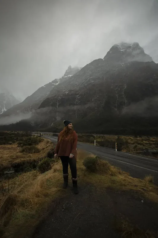 Monkey Creek: BEST Stop On The Way To Milford Sound 5 tasha amy wearing a rust-colored sweater and black pants stands near a winding road in Monkey Creek. The road leads towards dramatic, mist-covered mountains that tower in the background. The cloudy sky and rugged landscape create a sense of solitude and natural beauty.