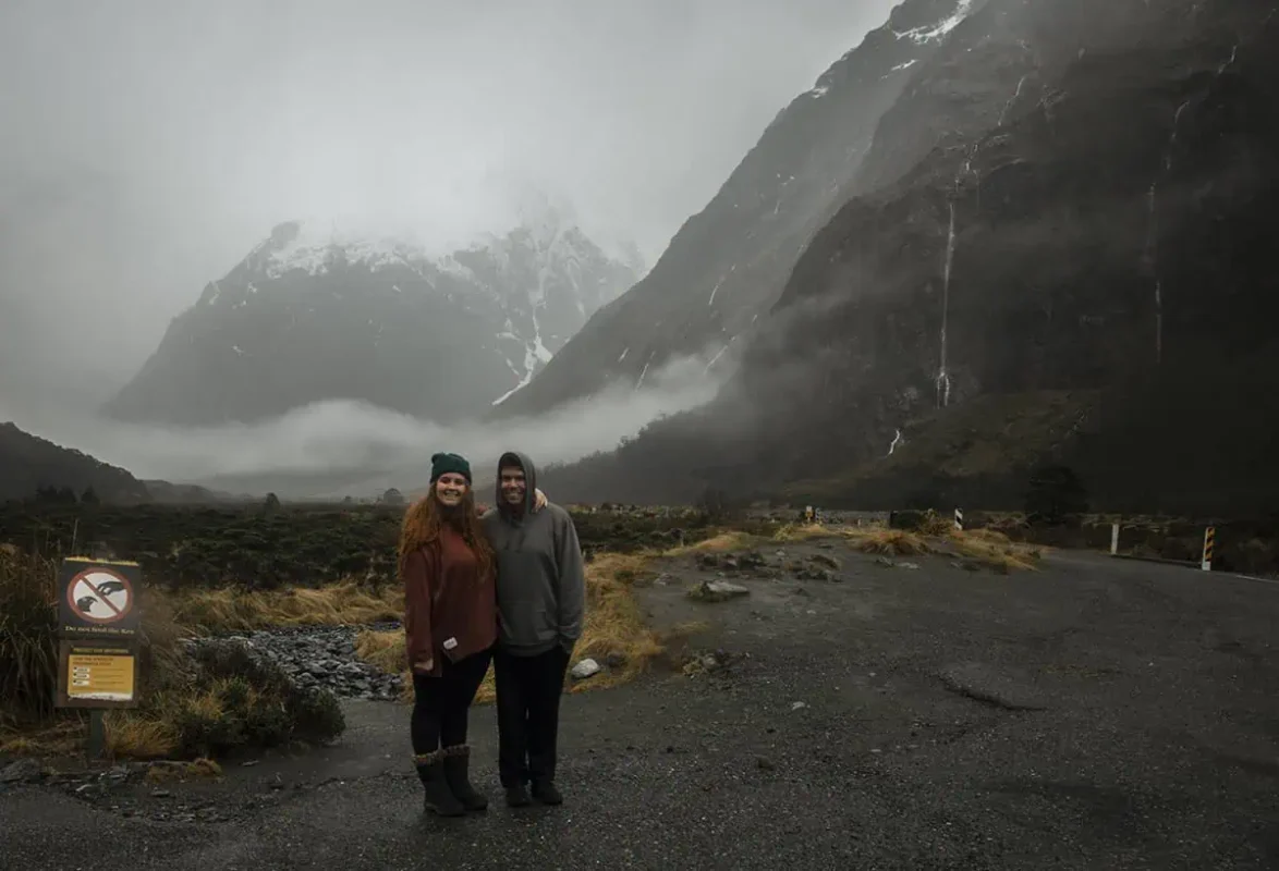 Monkey Creek: BEST Stop On The Way To Milford Sound 4 A couple poses together at Monkey Creek, New Zealand, with the woman wearing a rust-colored sweater and beanie and the man in a grey hoodie. They stand in front of a dramatic landscape of mist-covered mountains and a lush valley. A sign in the foreground advises against feeding the local kea birds, adding a touch of local wildlife awareness to the scene.
