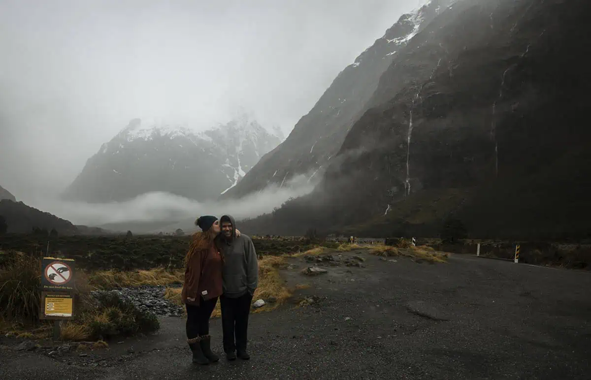 Monkey Creek: BEST Stop On The Way To Milford Sound 11 tasha amy and her partner james stand close together at Monkey Creek New Zealand. The woman kisses the man's cheek as they smile in front of the dramatic, mist-covered mountains. The overcast sky and rugged landscape provide a stunning natural backdrop, emphasizing the area's remote beauty