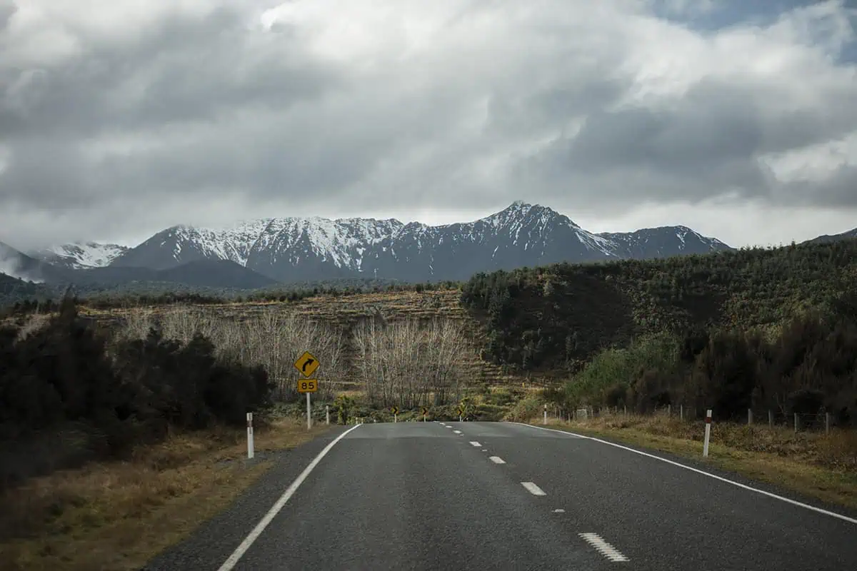 Queenstown To Milford Sound: Best Ways To Travel 2025 5 An open road leading towards snow-dusted mountain ranges under a cloudy sky. The sign on the side of the road indicates a sharp turn.