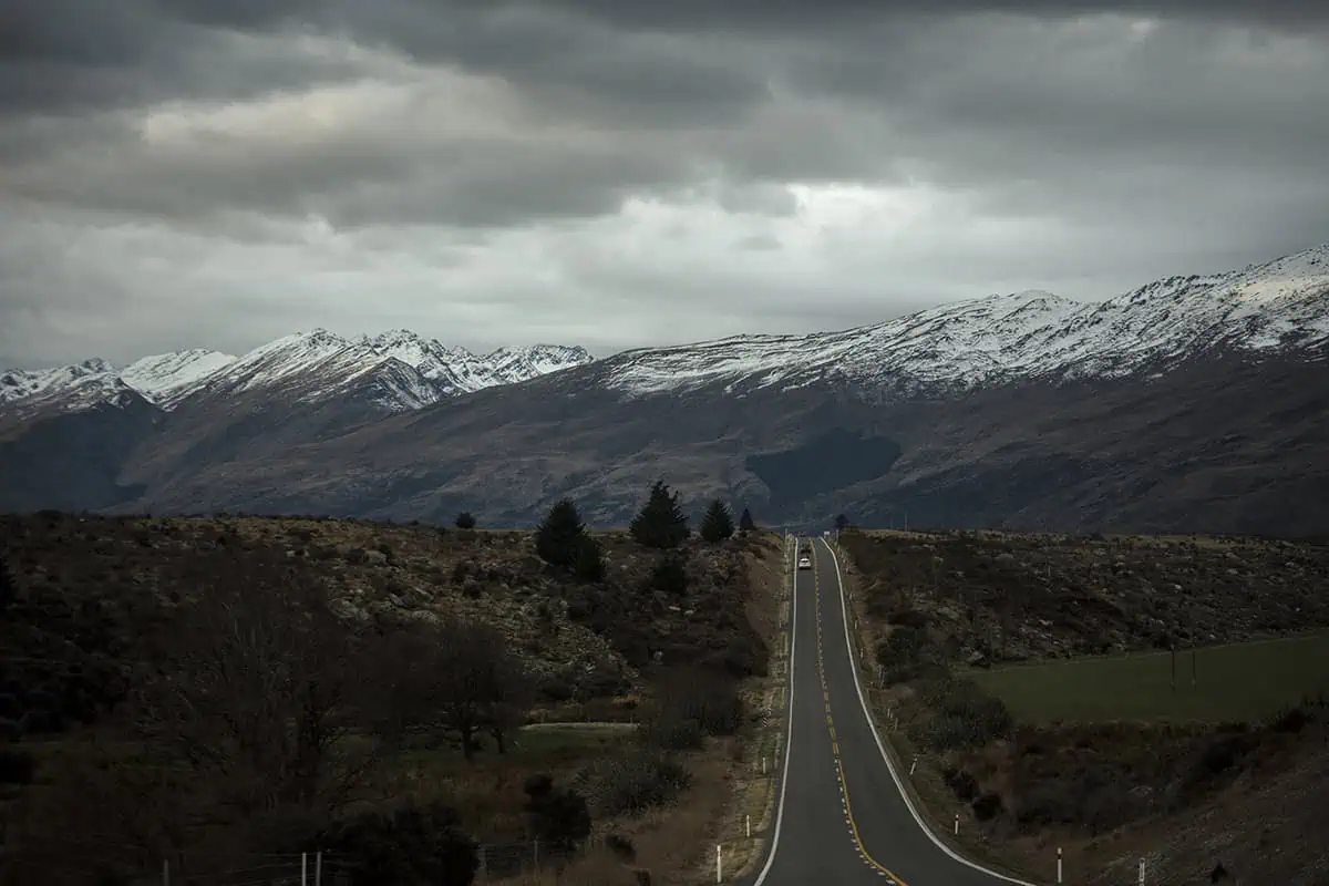 Queenstown To Milford Sound: Best Ways To Travel 2025 16 An open road stretches towards the distant, snow-capped mountains under a cloudy sky, with barren, rugged terrain on either side. This scene captures the remote and scenic drive from Queenstown to Milford Sound.