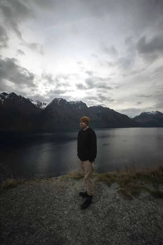 Queenstown To Milford Sound: Best Ways To Travel 2025 11 A person stands by the edge of a lake, with snow-capped mountains in the background under a cloudy sky. The reflective lake and towering peaks emphasize the tranquil yet majestic landscape encountered on the journey
