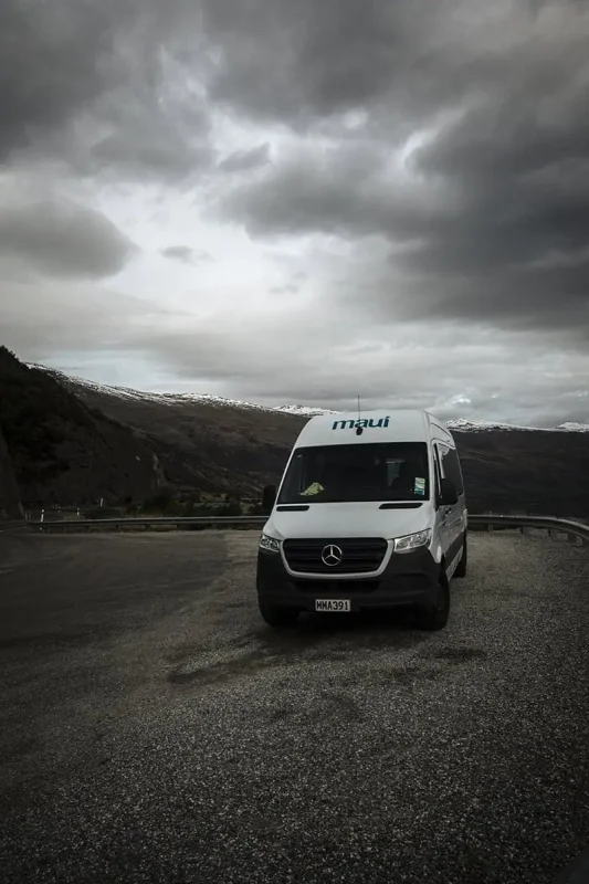 Queenstown To Milford Sound: Best Ways To Travel 2025 10 A white camper van marked "maui" is parked on a gravel pull-off area, with dramatic, overcast skies and mountain views in the background. The camper van represents the adventurous spirit of road-tripping from Queenstown to Milford Sound.
