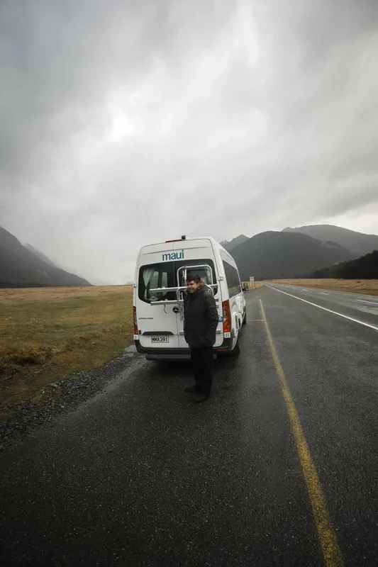 Queenstown To Milford Sound: Best Ways To Travel 2025 12 A white "maui" camper van is parked on the side of a wet road, with a person standing nearby in a dark coat. The misty, mountainous terrain and rainy conditions underscore the rugged journey from Queenstown to Milford Sound.