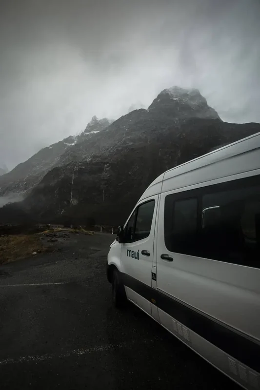 Queenstown To Milford Sound: Best Ways To Travel 2025 8 The white "maui" camper van is seen close-up, parked on a gravel area with towering, mist-covered mountains in the background. The dramatic scenery and overcast weather highlight the raw beauty of Milford Sound.