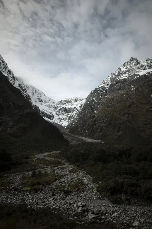 Queenstown To Milford Sound: Best Ways To Travel 2025 9 A view of a rocky, snow-covered mountain valley under a cloudy sky, with patches of forest and a dry riverbed below. The dramatic peaks and remote landscape emphasize the wild beauty of the Milford Sound region.