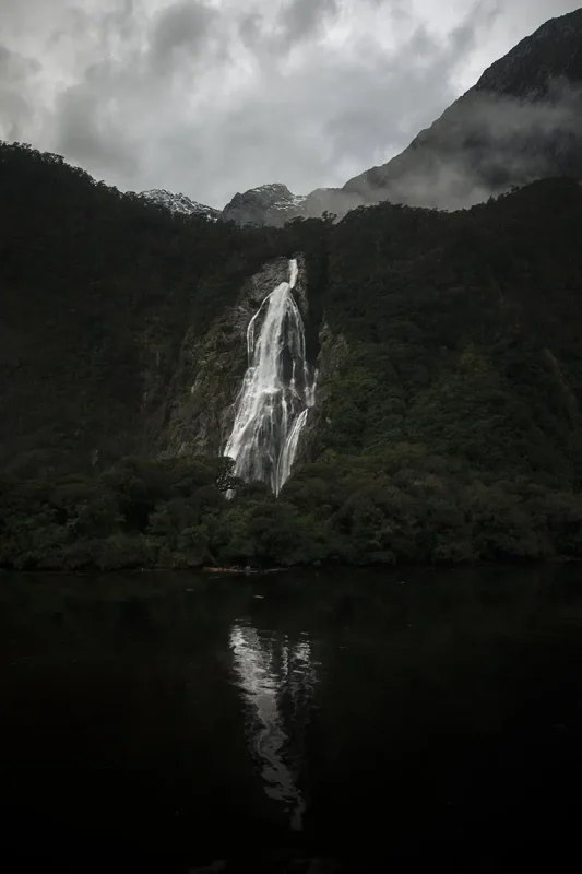 Queenstown To Milford Sound: Best Ways To Travel 2025 15 A powerful waterfall cascades down a lush, green mountainside into a dark, reflective lake. The mist from the falls and the cloudy sky above create a dramatic and picturesque view of Milford Sound.