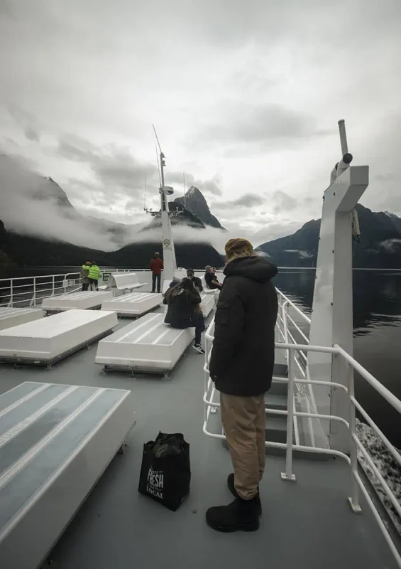 Queenstown To Milford Sound: Best Ways To Travel 2025 14 People stand on the deck of a boat, taking in the majestic views of Milford Sound's steep cliffs and cloudy sky.