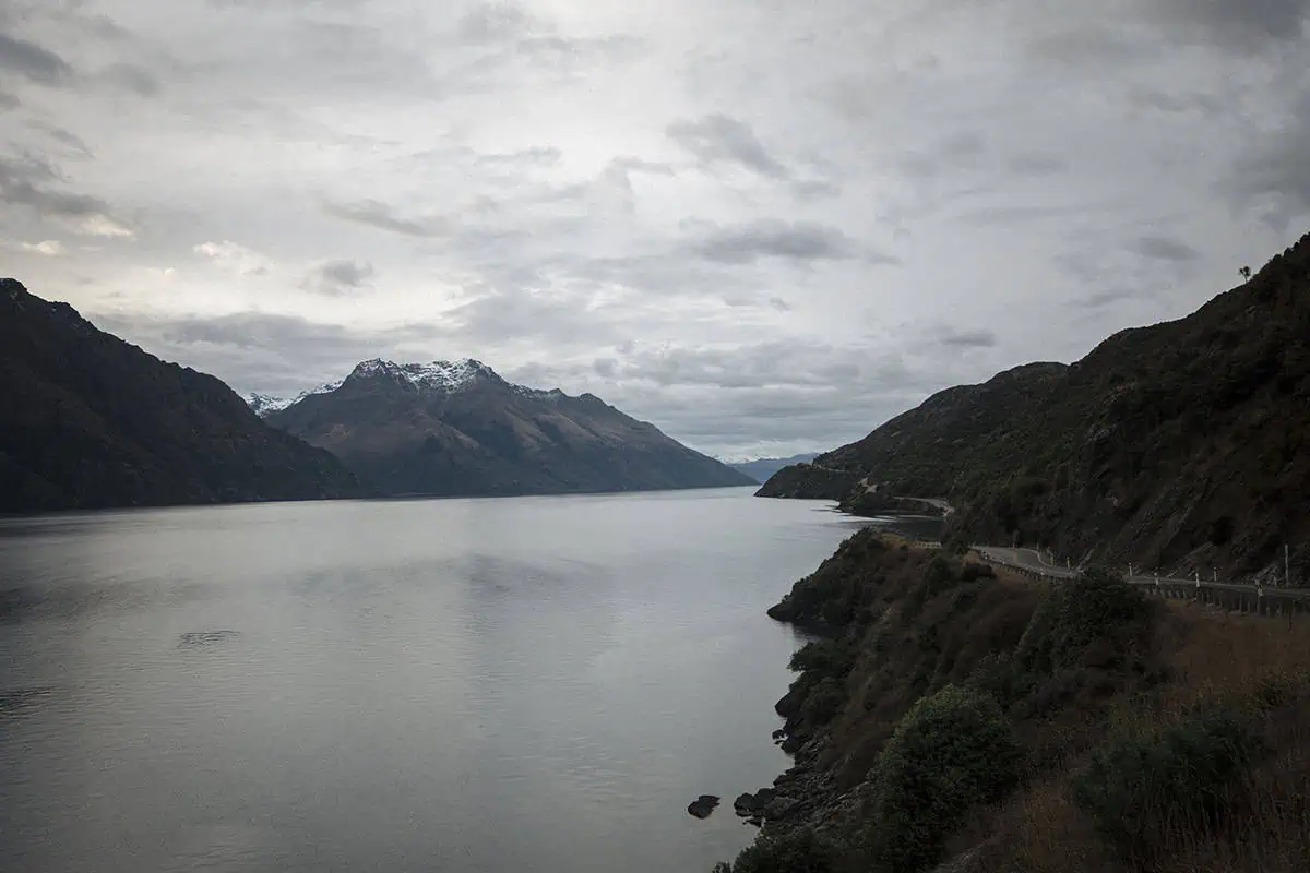 Queenstown To Milford Sound: Best Ways To Travel 2025 13 A serene view of Lake Wakatipu, with calm waters reflecting the overcast sky and snow-capped mountains in the background. The winding road along the lake hints at the journey from Queenstown to Milford Sound.