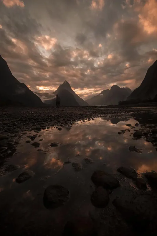 Milford Sound In The Rain: The BEST Time To Visit 2025 26 reflective pool with Milford Sound's iconic Mitre Peak in the background at sunset. The sky is ablaze with orange and pink hues, reflected in the still waters, creating a serene yet dramatic scene.