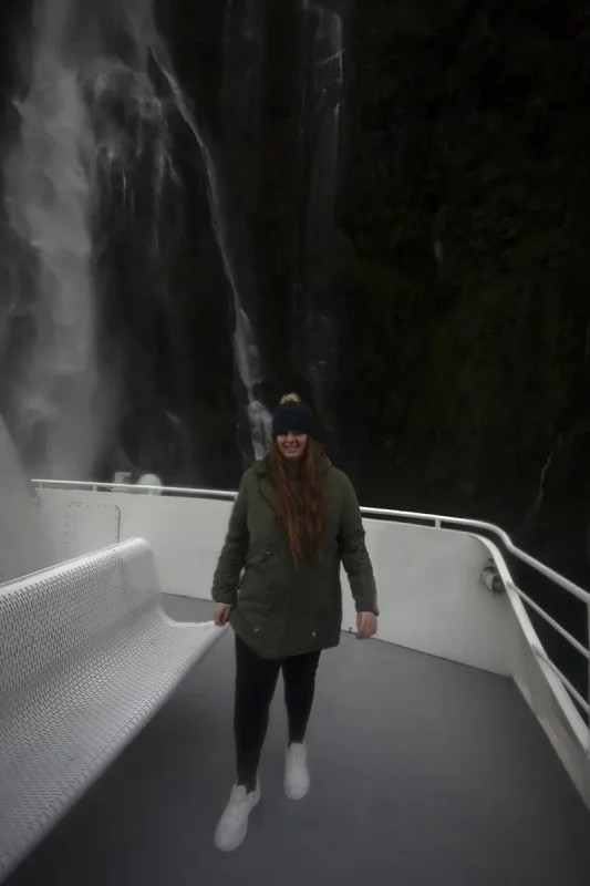 Milford Sound In The Rain: The BEST Time To Visit 2025 12 tasha amy in a green parka stands on a boat deck near a powerful waterfall in Milford Sound. The waterfall creates a misty spray, and the overcast sky adds to the sense of adventure and the typical wet weather of the region.