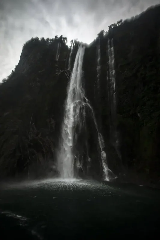 Milford Sound In The Rain: The BEST Time To Visit 2025 13 A majestic waterfall cascades down a sheer cliff into the dark waters of Milford Sound. The force of the water hitting the surface creates a dramatic splash, highlighted against the misty and overcast conditions.