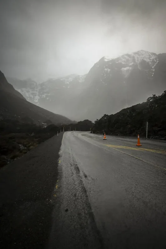 Milford Sound In The Rain: The BEST Time To Visit 2025 18 A wet road winds through a misty mountain landscape in Milford Sound. The road is lined with orange traffic cones, and the overcast sky and low-hanging clouds create a moody and serene atmosphere.