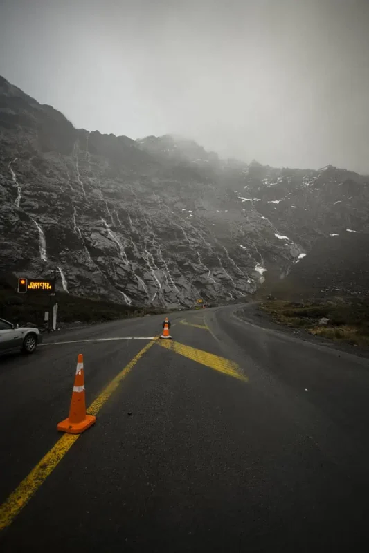 Milford Sound In The Rain: The BEST Time To Visit 2025 17 A rain-soaked road curves around a rocky mountainside with thin waterfalls cascading down in Milford Sound. Orange traffic cones and a warning sign are visible, emphasizing the rugged and wet conditions of the area.