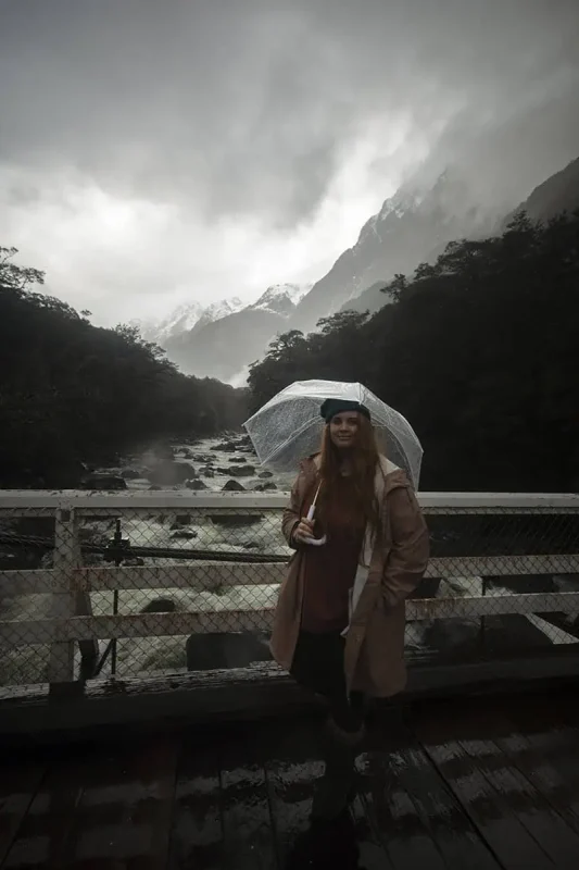 Milford Sound In The Rain: The BEST Time To Visit 2025 10 tasha amy stands on a bridge holding an umbrella, overlooking a fast-flowing river surrounded by misty mountains in Milford Sound. The overcast sky and low clouds highlight the typical rainy weather in the region.