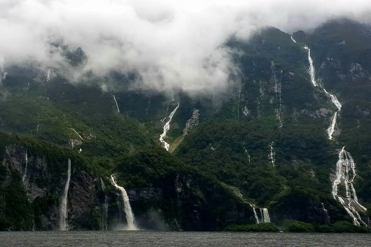 Milford Sound In The Rain: The BEST Time To Visit 2025 8 aterfalls cascade down lush, green mountains shrouded in mist and low-hanging clouds in Milford Sound. The dramatic scenery captures the raw beauty of the landscape, with multiple waterfalls visible against the dense foliage. This image perfectly showcases the ethereal atmosphere of rain in Milford Sound.