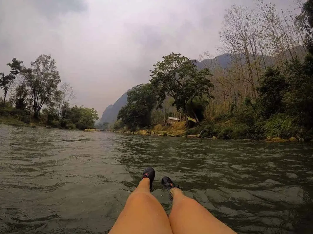 Tubing In Vang Vieng, Laos: ULTIMATE GUIDE 2025 9 Person's perspective of their legs floating on a river with lush greenery and mountains in the background, capturing the essence of tubing in Vang Vieng