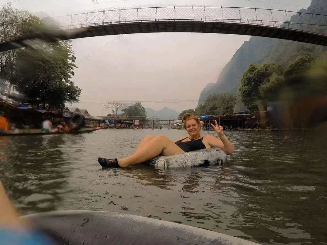 Tubing In Vang Vieng, Laos: ULTIMATE GUIDE 2025 12 Tasha Amy smiling and making a peace sign while floating on a tube in a river under a bridge with a rustic village backdrop in Laos