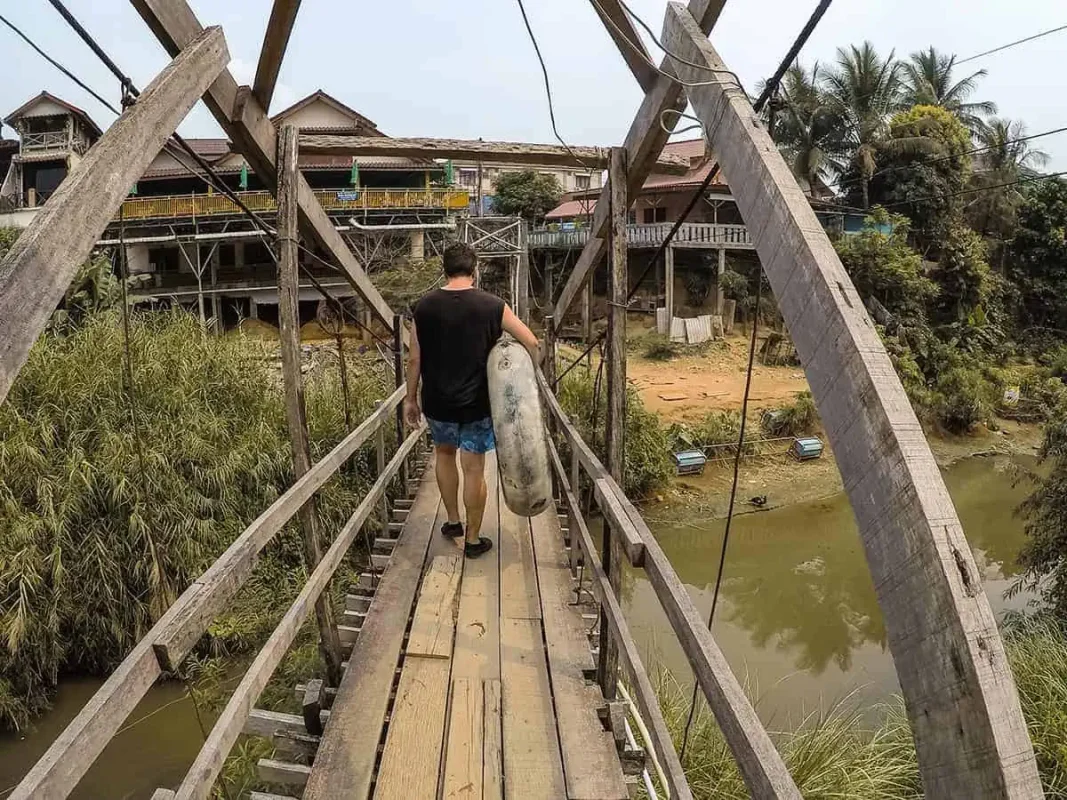 Tubing In Vang Vieng, Laos: ULTIMATE GUIDE 2025 13 A person walking on a wooden bridge carrying a tube, with traditional houses on stilts and dense vegetation on either side.