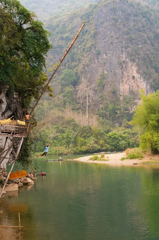 Tubing In Vang Vieng, Laos: ULTIMATE GUIDE 2025 6 A daring individual swings from a high platform on a rope swing above a tranquil river surrounded by steep cliffs and greenery in Laos.