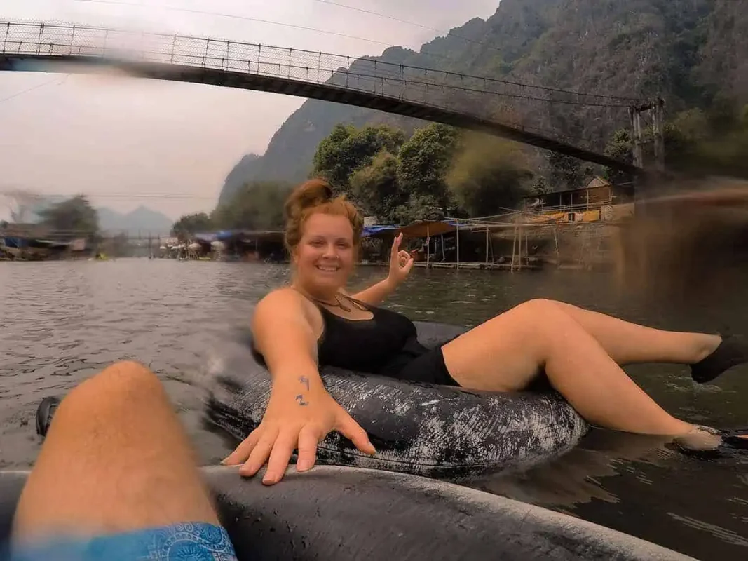 Tubing In Vang Vieng, Laos: ULTIMATE GUIDE 2025 7 Tasha Amy n a black outfit floats on a tube in a murky river, waving at the camera with a bridge and village in the background.