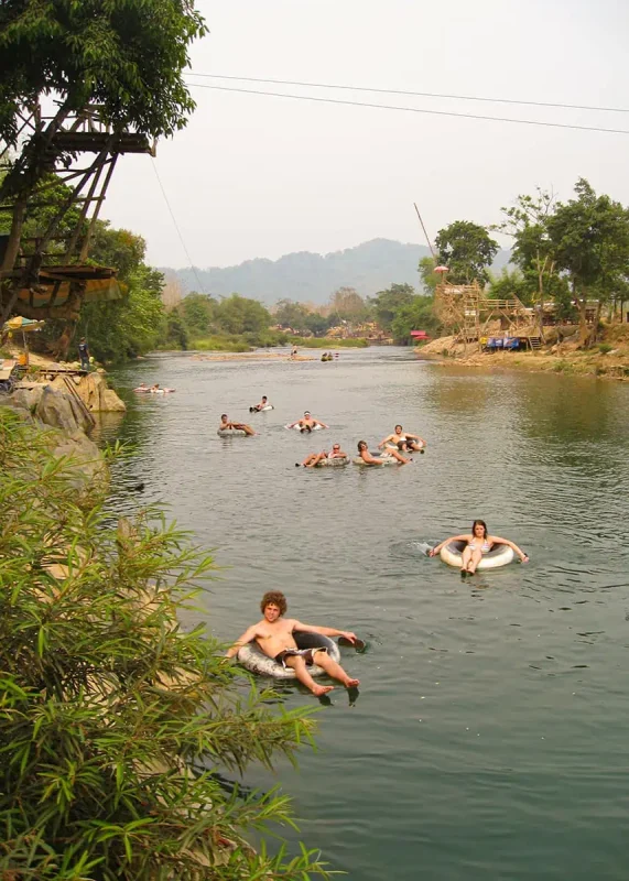 Tubing In Vang Vieng, Laos: ULTIMATE GUIDE 2025 5 A serene river scene with people tubing, a wooden platform with onlookers, and a lush riverside landscape in Laos.