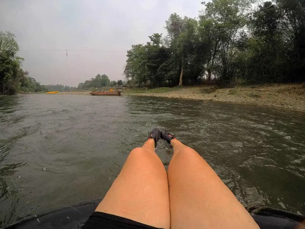 Tubing In Vang Vieng, Laos: ULTIMATE GUIDE 2025 11 A first-person view of a person's feet extended over a black inner tube, floating down a murky river with trees lining the shore and a yellow kayak in the distance, a typical scene of tubing in Vang Vieng.