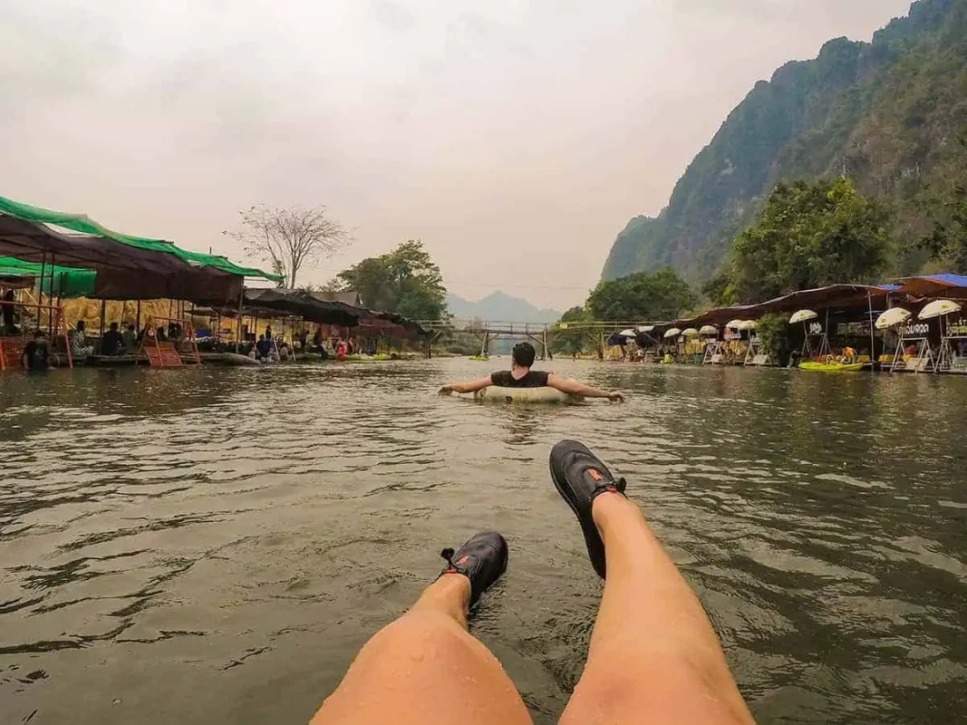 Tubing In Vang Vieng, Laos: ULTIMATE GUIDE 2025 4 A person's legs seen from their perspective while floating on a tube in a river, following another person in the water, with a row of riverside huts and mountain scenery in the background, capturing the relaxed atmosphere of tubing in Laos.
