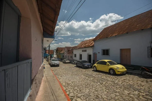 A cobblestone street lined with colorful buildings and parked cars after arriving from San Ignacio and Flores. The bright yellow VW Beetle stands out under the clear sky.