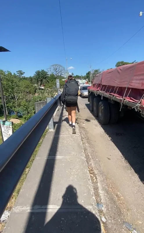 Travel San Ignacio To Flores & Crossing The Border 22 A female backpacker with a large black backpack and wearing a black tank top, shorts, and white socks walks on a narrow sidewalk beside a parked red tarp-covered truck when traveling from san ignacio to flores. The sidewalk is raised, adjacent to a guardrail overlooking a river, and leads through a small town with colorful buildings and clear skies.