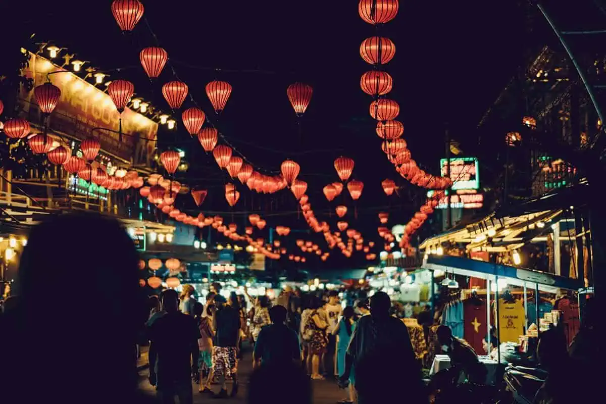 Scams In Vietnam: READ BEFORE YOU VISIT (2025) 8 A busy night market in Hoi An, Vietnam lit by red lanterns. There's a crowd of people shopping the colorful stalls.