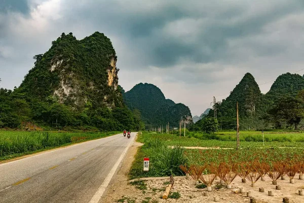 A road in Vietnam with motorbikes riding through and limestone mountains under a cloudy sky. A roadside milestone labeled H7 302 marks the route, and possible scams in vietnam.