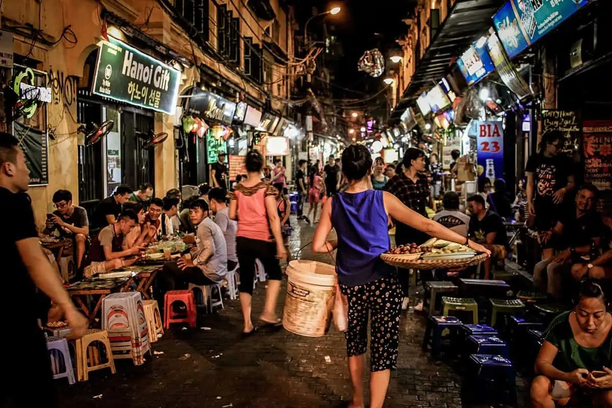 Scams In Vietnam: READ BEFORE YOU VISIT (2025) 13 Crowded night street in Vietnam’s Old Quarter, where tourists and locals dine on plastic stools while street vendors walk between the tables selling snacks.