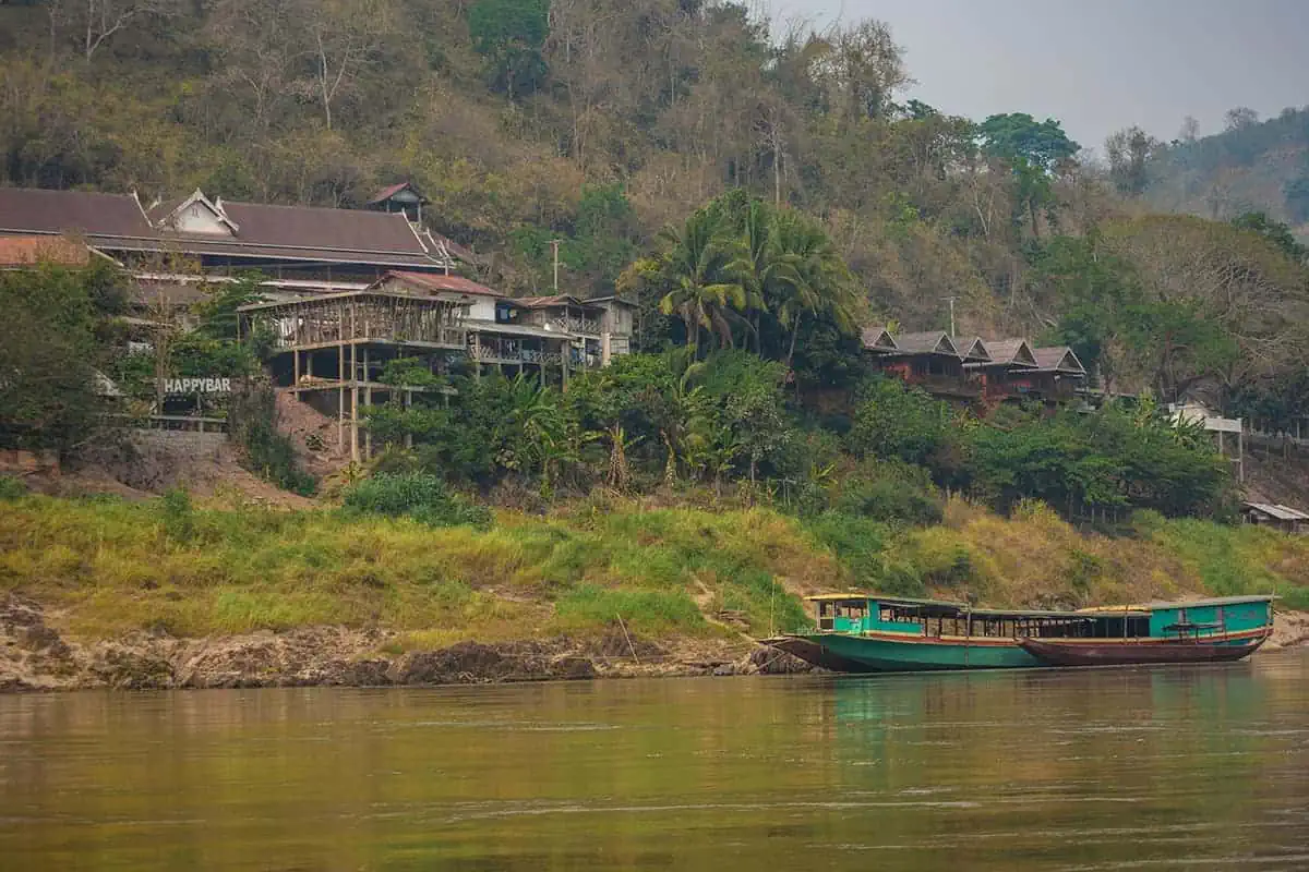 Taking The Slow Boat To Luang Prabang: BEST Tips 2025 13 A boat cruises by a lush riverbank with traditional Lao architecture in a village called Pakbeng