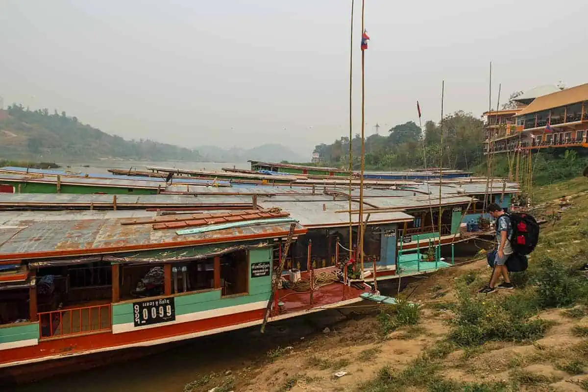 Chiang Mai To Luang Prabang: All You Need To Know 2025 13 Travelers with backpacks approach a fleet of slow boats docked at the riverbank