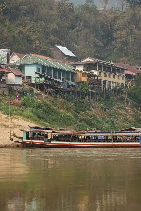 Taking The Slow Boat To Luang Prabang: BEST Tips 2025 15 A slow boat on the river against a backdrop of colorful houses.