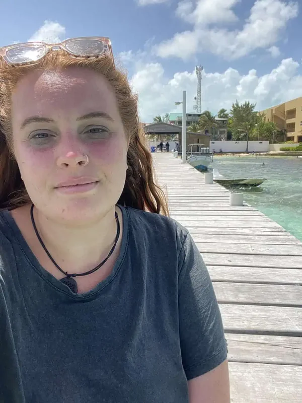 Solo Travel In Belize: My Experience As A Solo Female Backpacker 9 A close-up selfie of tasha amy with auburn hair, wearing a grey T-shirt and sunglasses perched on her head, standing on a long wooden pier that stretches into the turquoise sea. The background features a mix of beachfront buildings and swaying palm trees under a bright sky. Her slight smile and sun-kissed face reflect the joy and relaxation of solo travel in Belize.