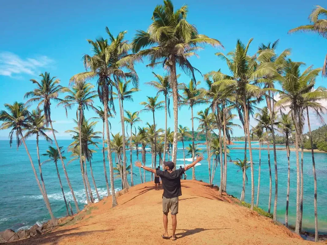 22+ Cheapest Countries To Backpack In The World! (2025) 2 A backpacker standing on a dirt road with palm trees in the background, surrounded by the beauty of a backpacker-friendly country of sri lanka