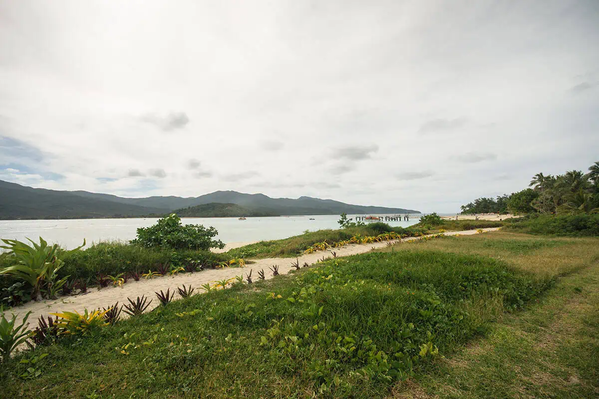 Things To Do On Mystery Island In Vanuatu: For Cruise Passengers & More 9 Looking across a grassy patch and sandy trail on Mystery Island toward a wooden pier and the mountainous coastline across the water.