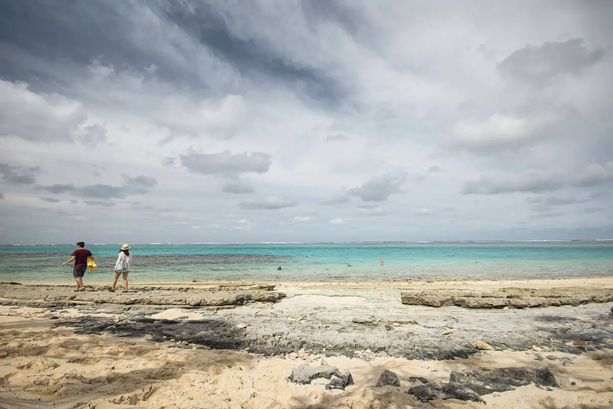 Things To Do On Mystery Island In Vanuatu: For Cruise Passengers & More 10 Two people walk along the rocky beach of Mystery Island under a dramatic, cloud-filled sky, while others swim in the shallow turquoise lagoon.
