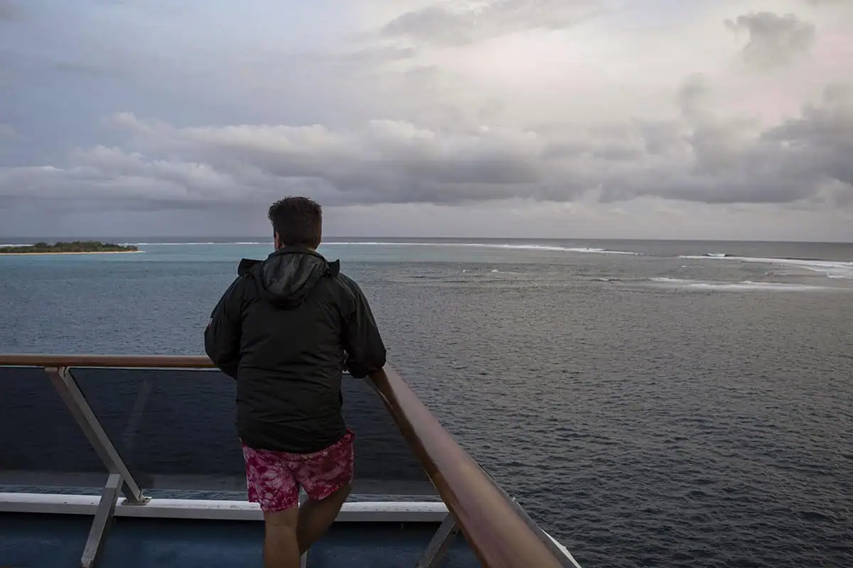 Things To Do On Mystery Island In Vanuatu: For Cruise Passengers & More 22 A man in a black jacket and red swim trunks stands on a cruise ship deck, gazing at Mystery Island across turquoise waters under a moody, overcast sky.