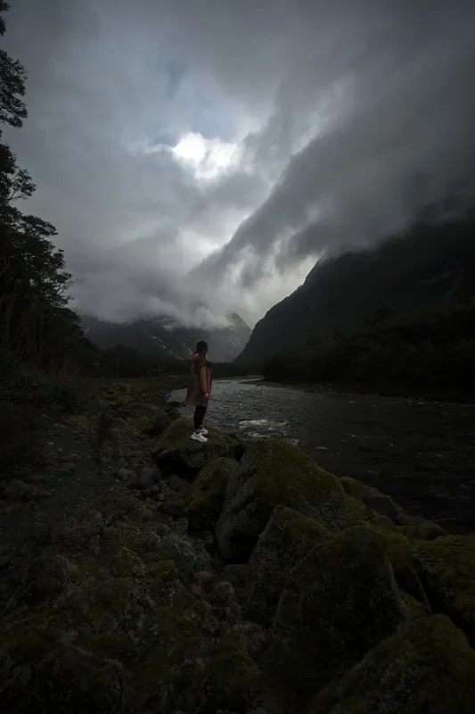 Best Waterfalls In Milford Sound Worth Checking Out 21 tasha amy n a raincoat stands on mossy rocks beside a river, gazing at the mist and cliffs of Milford Sound.