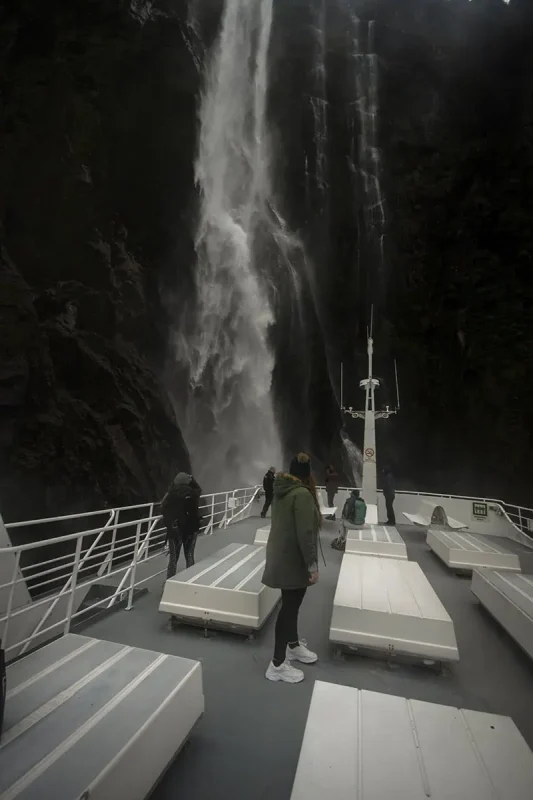 Best Waterfalls In Milford Sound Worth Checking Out 7 Visitors on the upper deck of a cruise boat stand in awe beneath a powerful stirling falls crashing down directly in front of them in Milford Sound.