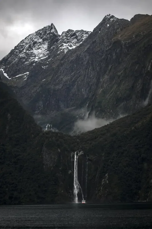 Best Waterfalls In Milford Sound Worth Checking Out 9 A dramatic waterfall plunges from a forest into the fiord below, with a small cruise boat at its base and snow-dusted peaks behind.