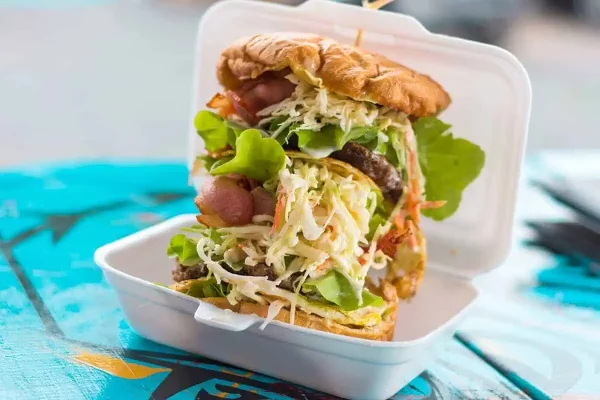 Famous Palace Takeaways burger, a budget-friendly option when exploring where to eat in Rarotonga. An overstuffed takeaway burger packed with grilled meat, lettuce, and sauce in a styrofoam container, a popular choice at Rarotonga restaurants.