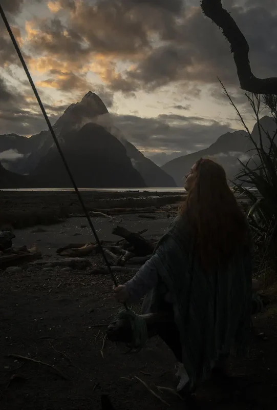 Milford Sound In Winter: Is It Still Worth The Hype? 21 tasha amy sits on a rustic swing made of a wooden log and ropes, set against the backdrop of Milford Sound at dusk. The sky is painted with vibrant shades of orange and purple, reflecting on the calm water. The mist-covered peaks add to the dramatic and serene atmosphere of winter in Milford Sound.